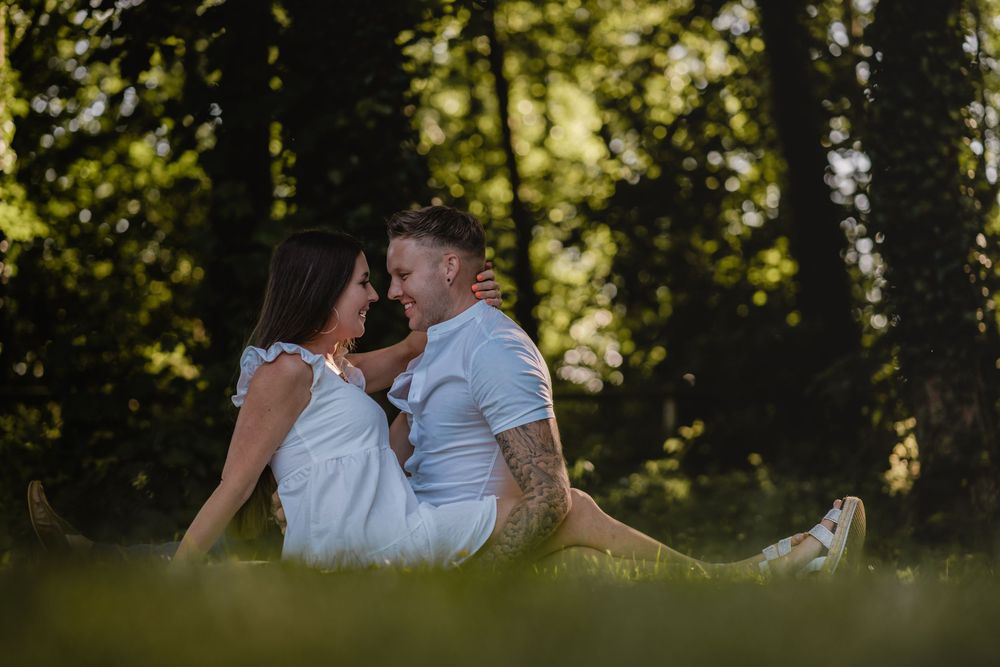 Romantic couple sitting close together on grass with forest backdrop.