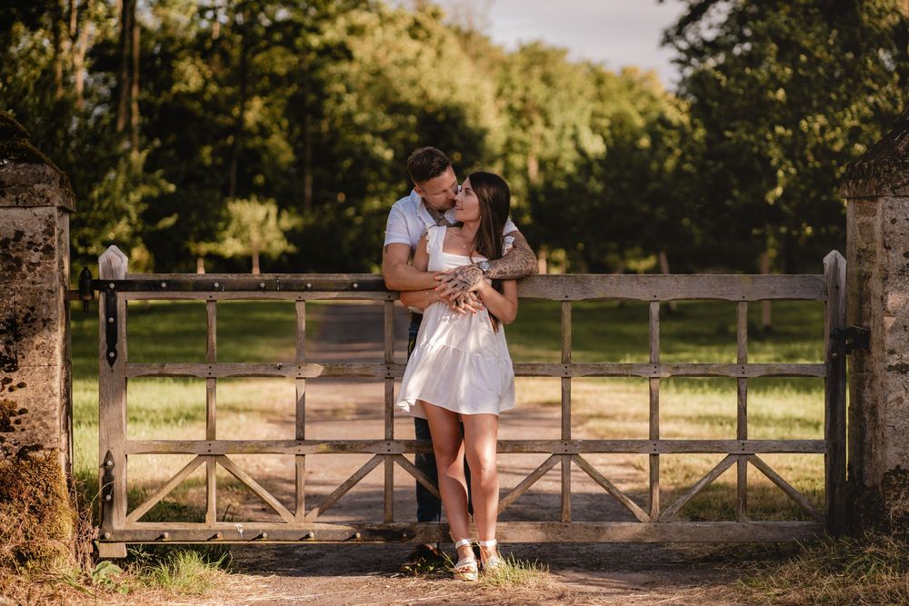 A tender moment between a couple leaning against a wooden gate in a park setting.