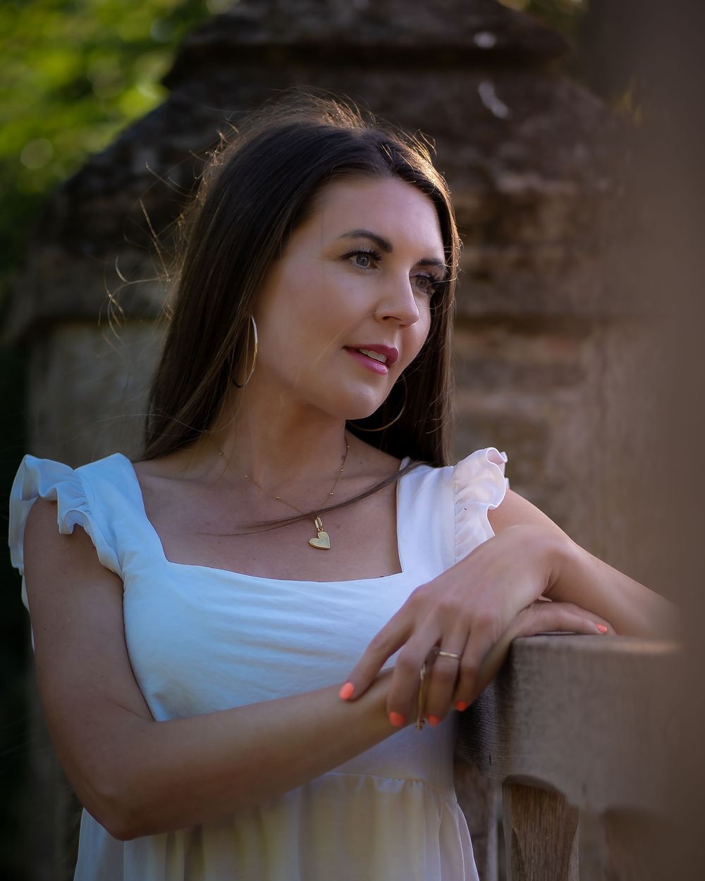 Woman in white ruffled dress poses against stone wall with golden sunlight in background.