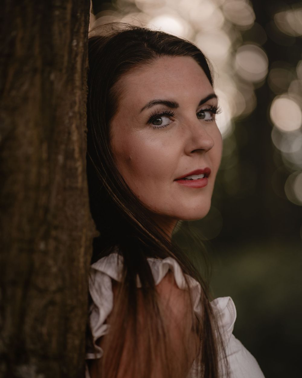 Portrait photo with warm bokeh background showing a woman in white blouse next to a tree.