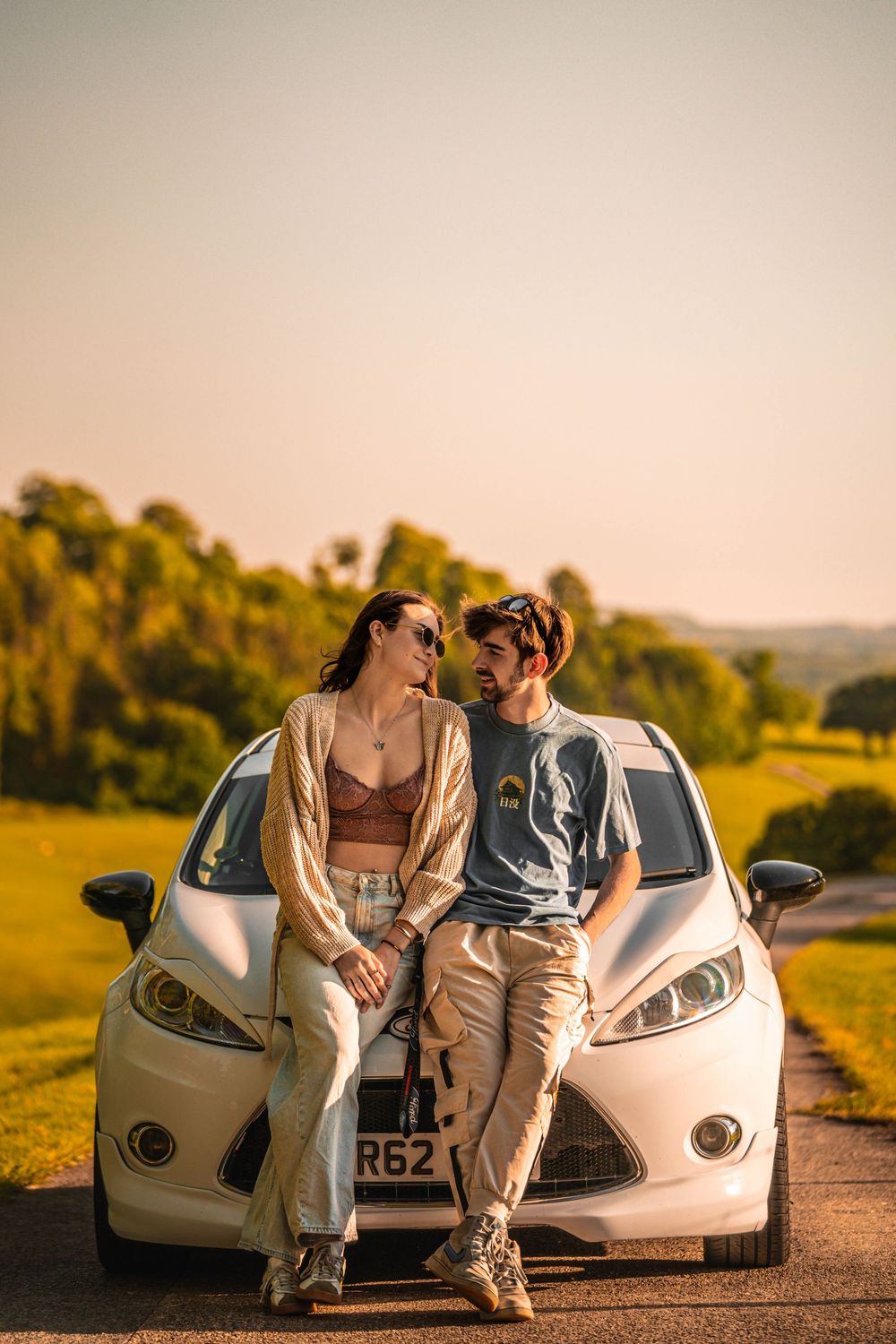 Two people sitting on the hood of a white Ford Fiesta during golden hour.