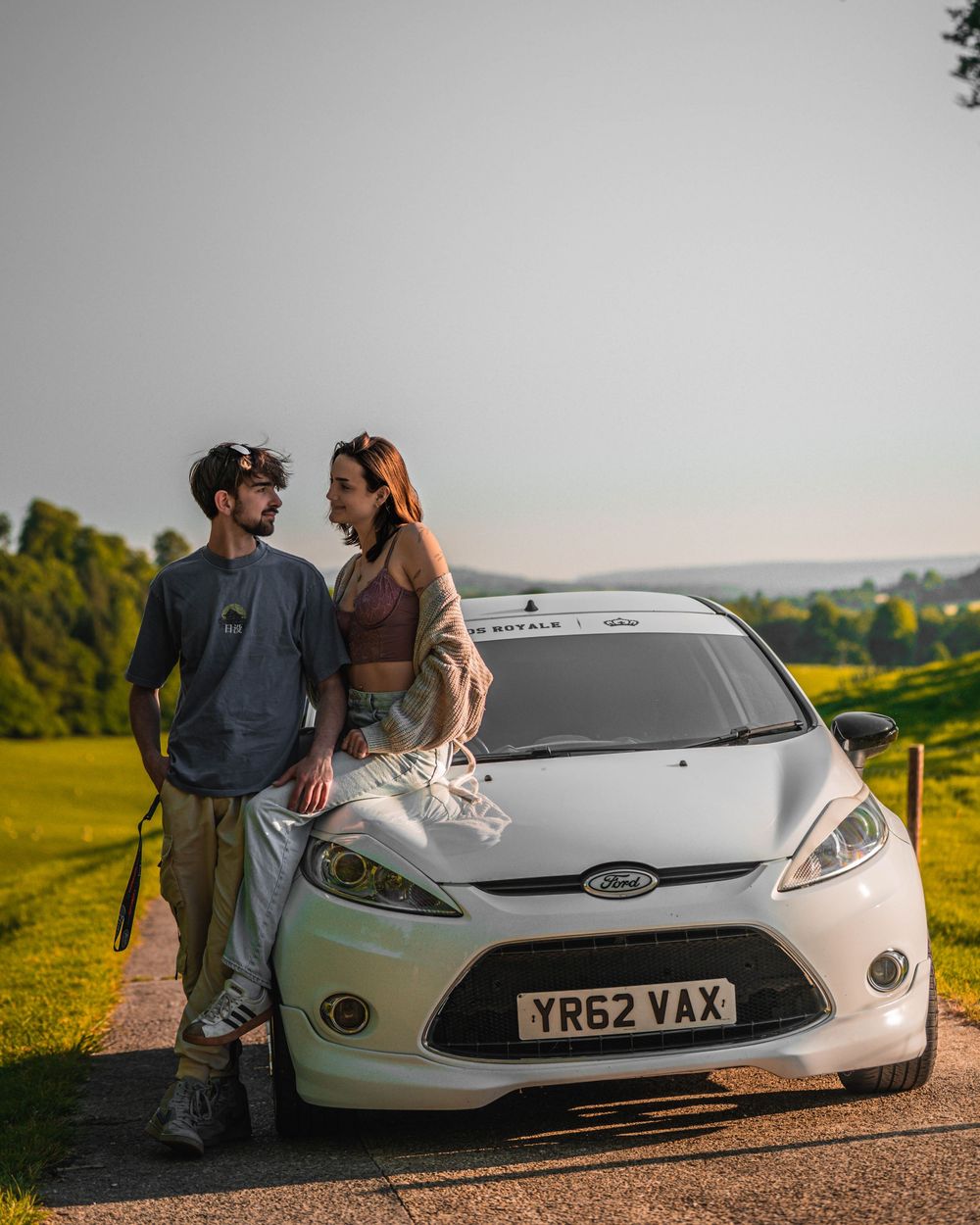 A couple poses with their white Ford Fiesta on a hilltop during sunset.