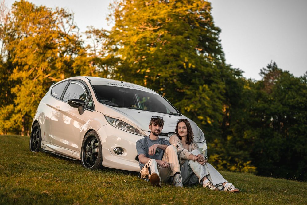 A couple sits by their modified white Ford Fiesta against autumn trees.
