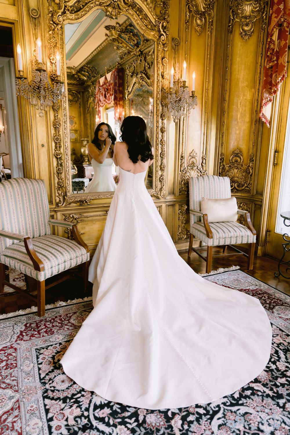 Bride in elegant white ball gown with train stands before ornate gilded mirror in luxurious baroque room.