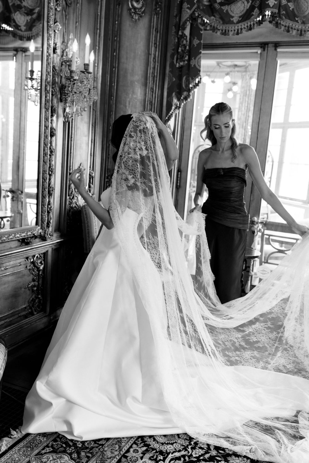Black and white photo of a bride in a lace veil and gown with a bridesmaid in a strapless dress in an ornate room.