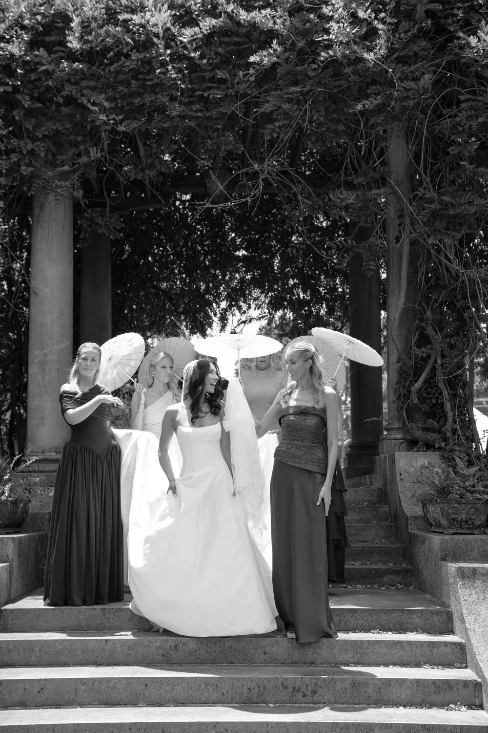 Black and white photo of a bride with three bridesmaids holding parasols on stone steps under a garden pergola.