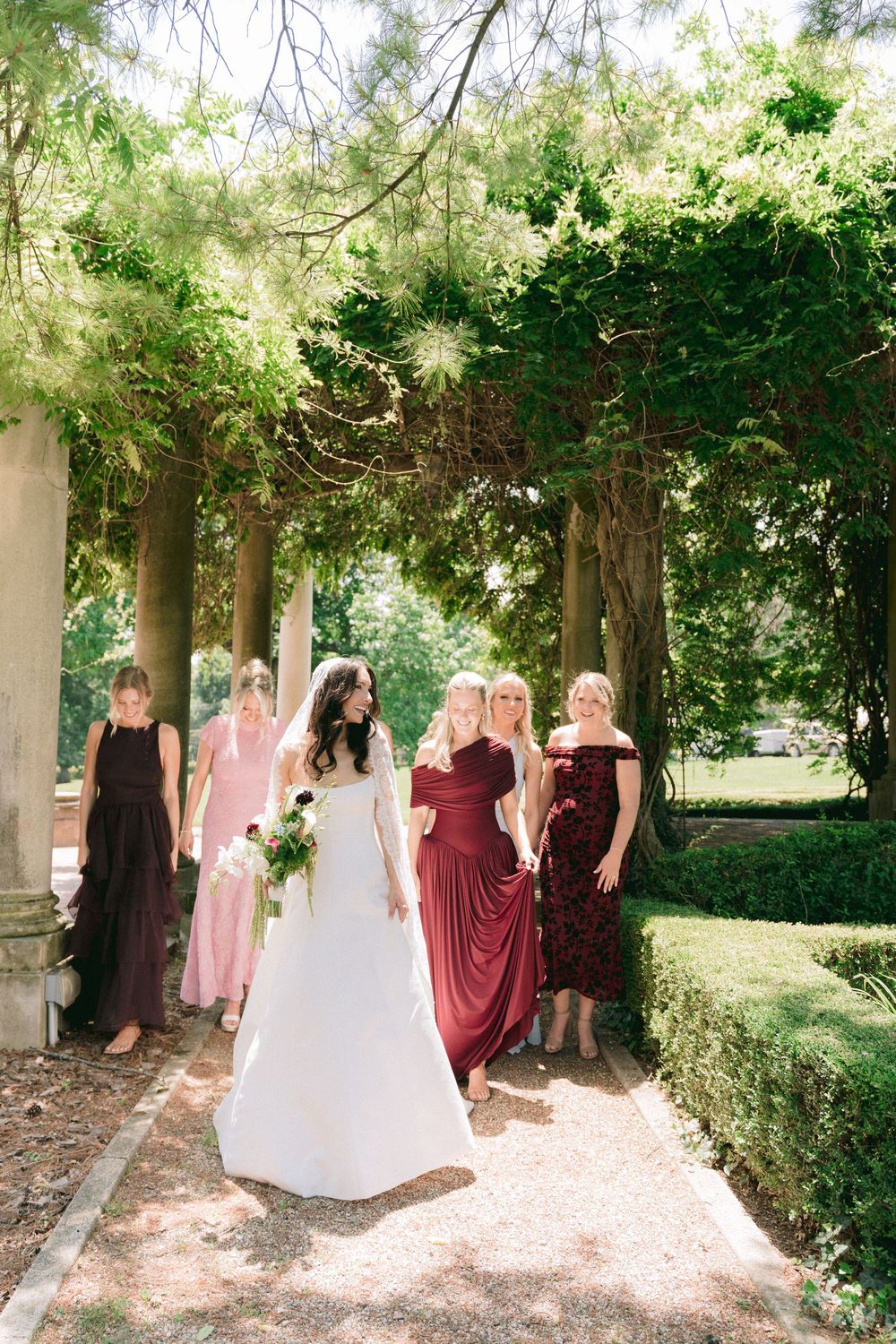Bride in white gown walks with bridesmaids in burgundy and pink dresses along a tree-lined garden path.