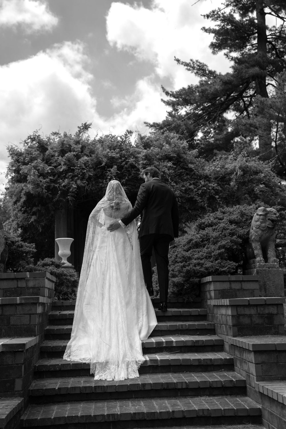 Black and white photo of bride and groom walking up stone steps in a lush garden on their wedding day.