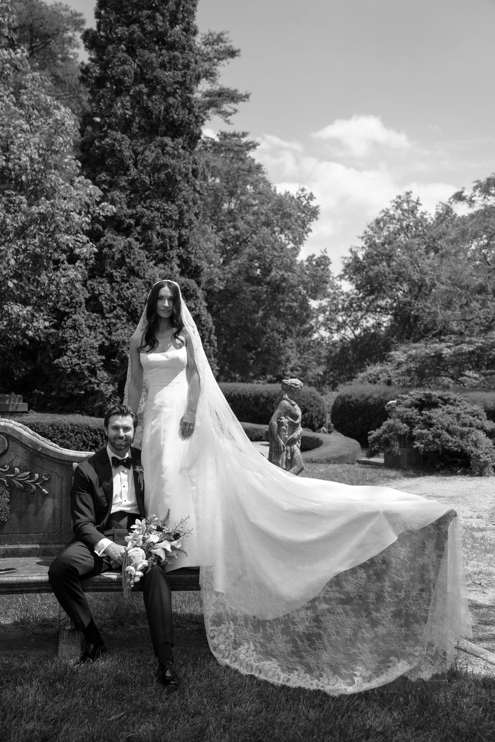 Black and white wedding photo of bride in flowing gown and veil with groom in tuxedo seated on bench in garden.