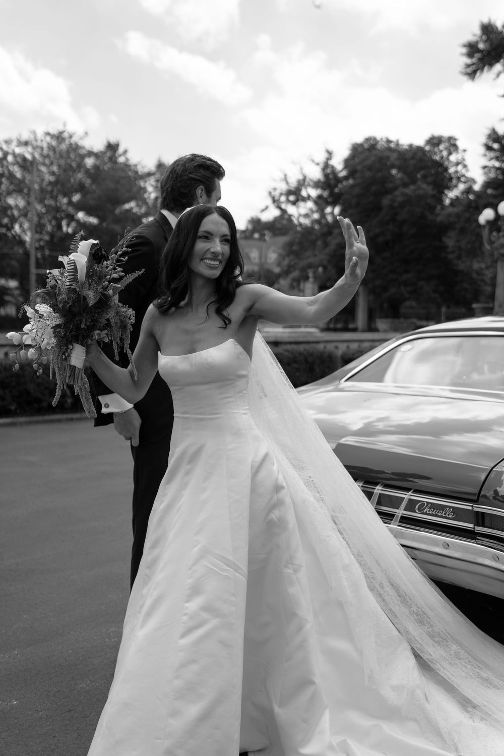 Smiling bride in strapless ball gown waves while holding bouquet beside vintage car in black and white photo.