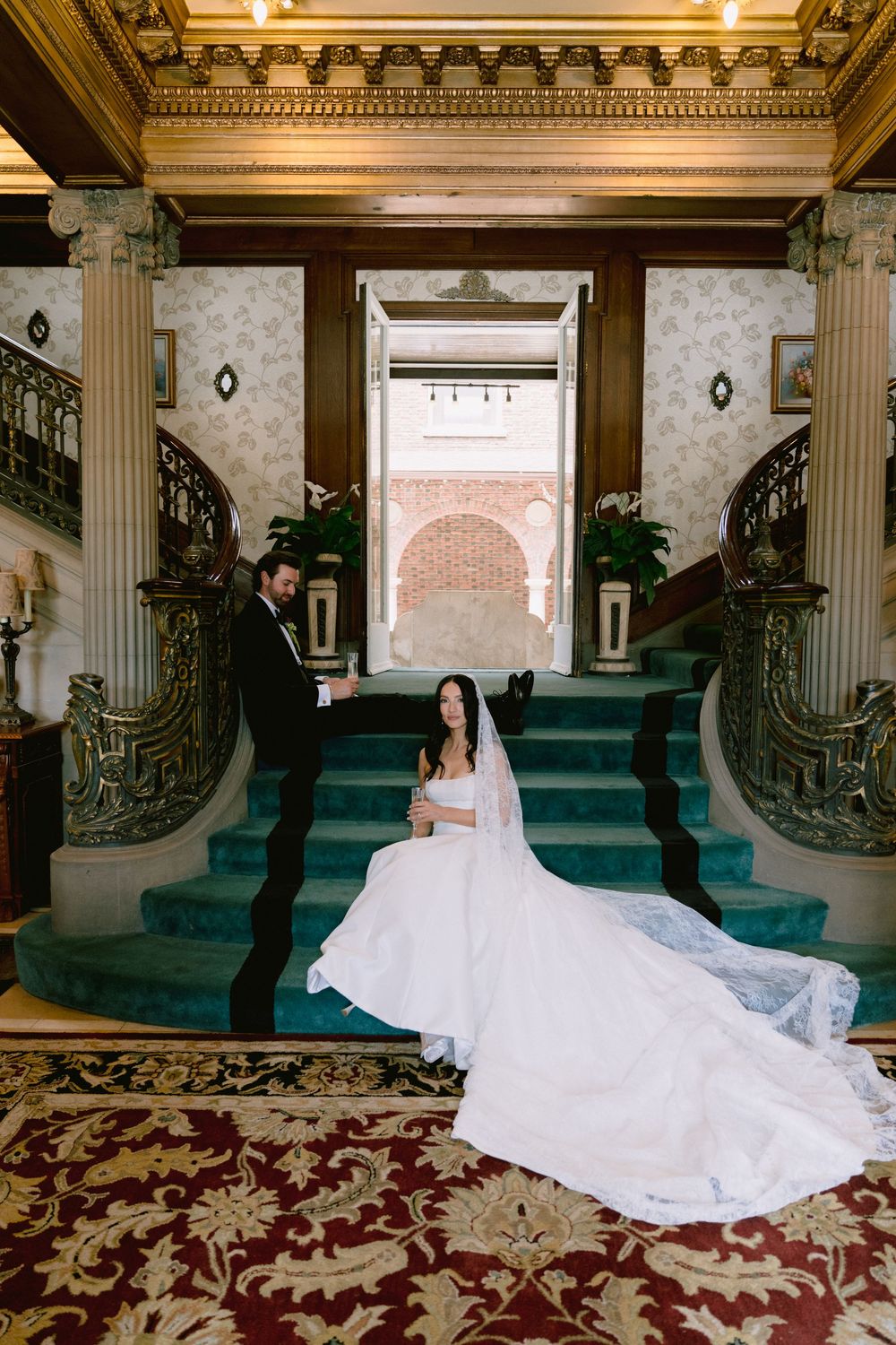 Bride in white gown sits on green carpeted staircase with groom in tuxedo inside elegant historic mansion.