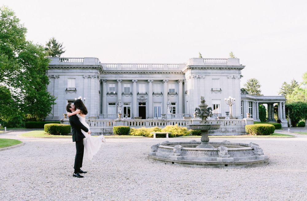 Couple embracing in front of elegant neoclassical mansion with stone fountain during outdoor wedding photo session.