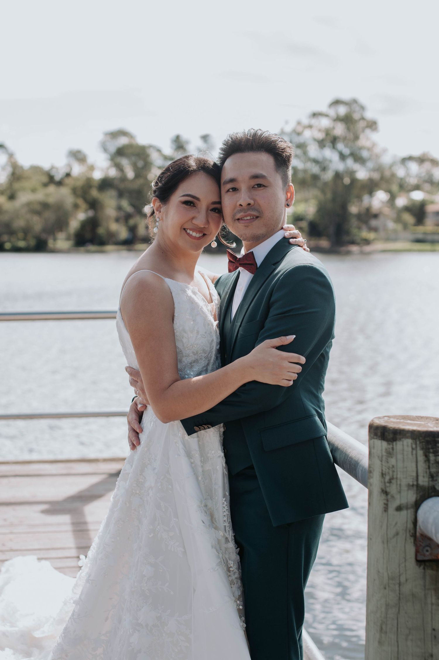 A couple in wedding attire embrace on a wooden dock by a serene lake with trees in the background.