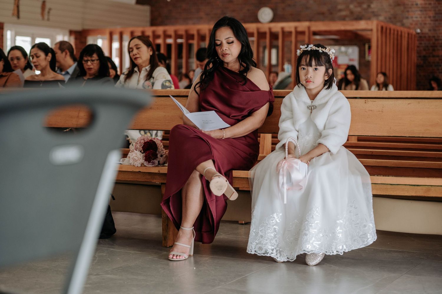 People sitting in wooden church pews during a formal ceremony, with a woman in maroon and child in white dress.