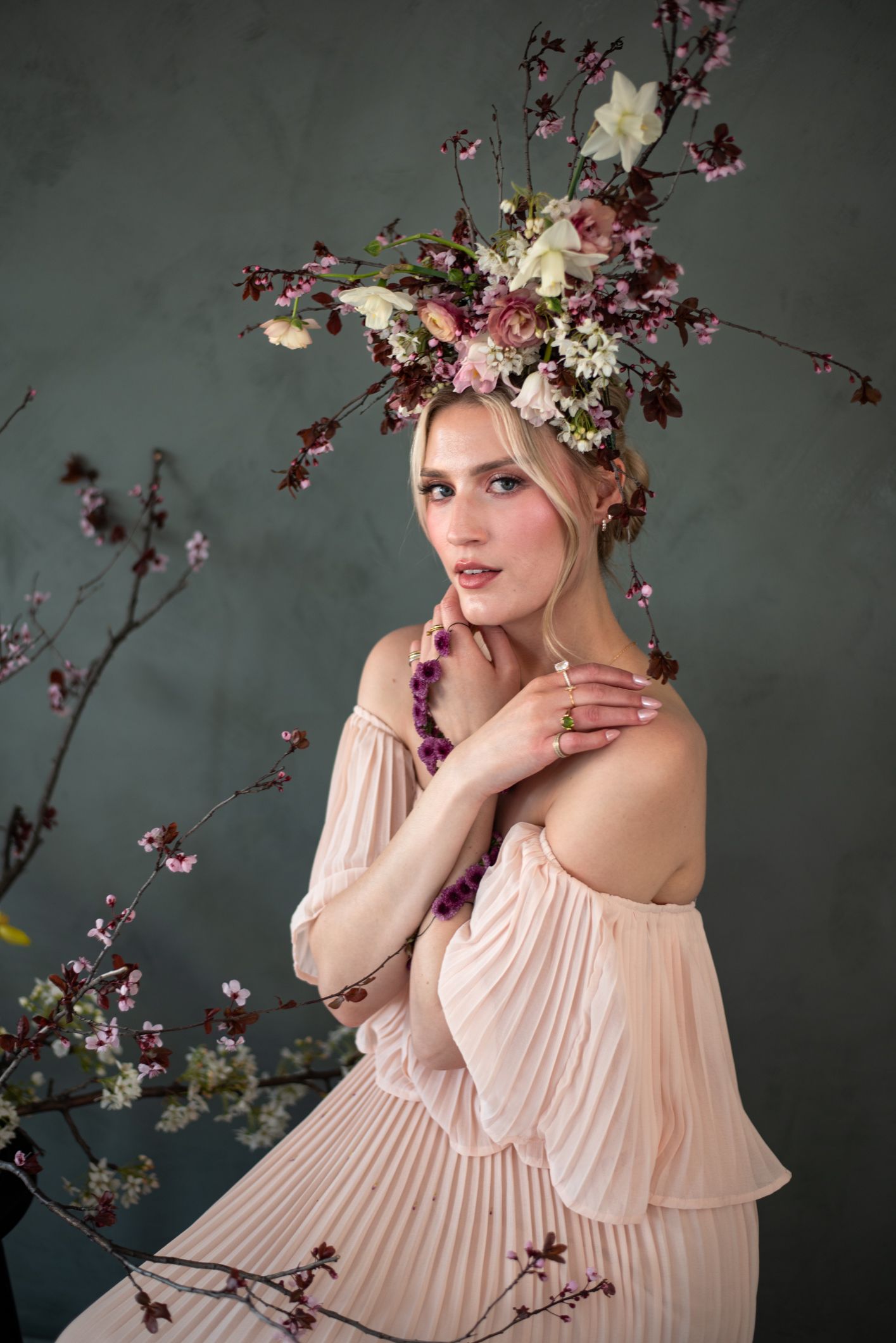 A whimsical spring portrait featuring a floral crown of white lilies and purple blooms over pink tulle dress against gray backdrop.