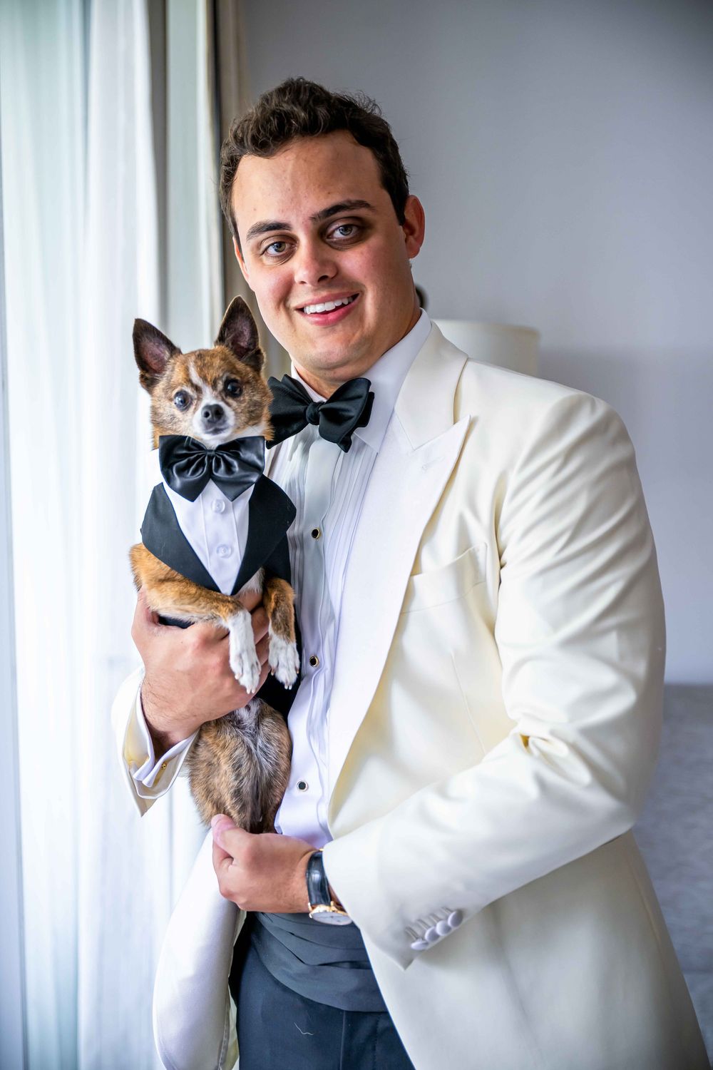 A person in a white tuxedo jacket holds a small dog wearing a matching bowtie and tuxedo outfit.