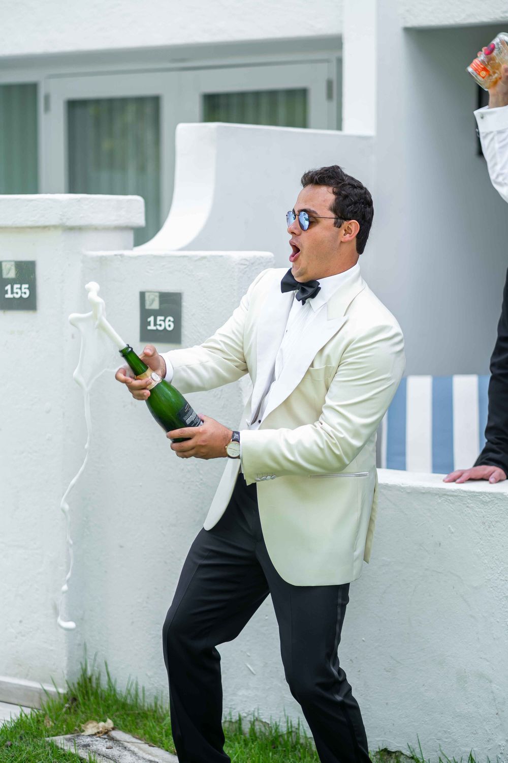 A person in a white tuxedo jacket and black pants pops open a champagne bottle with excitement outside.