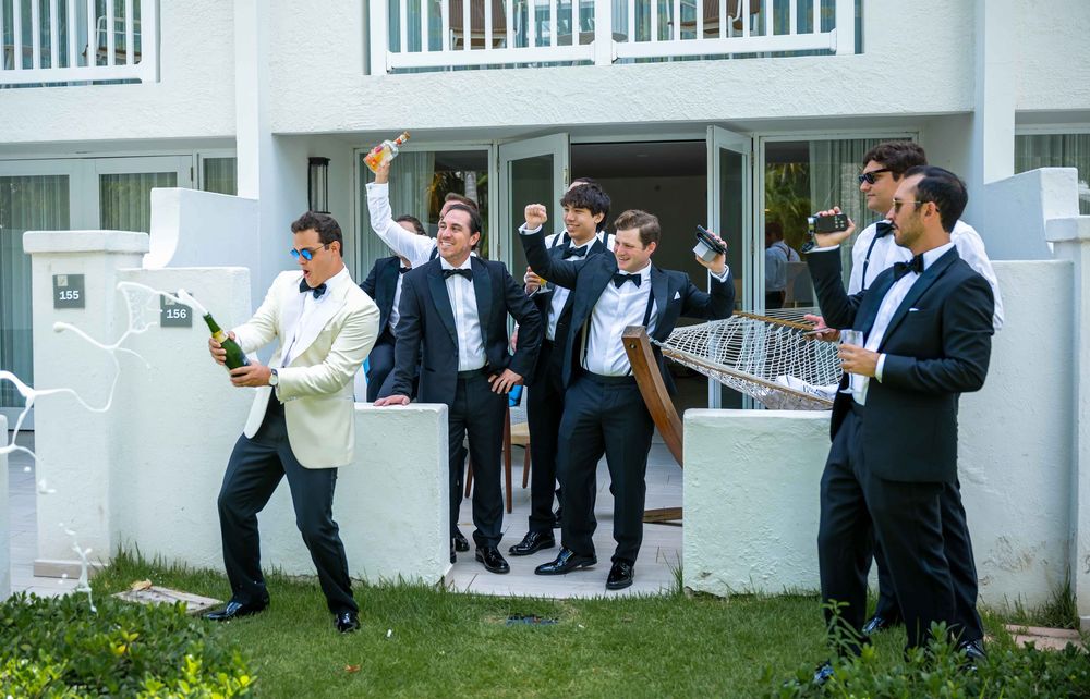 Groomsmen in formal attire pose together and celebrate outside while holding beverages at an outdoor venue.