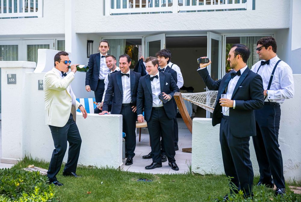 Wedding party groomsmen in formal tuxedos and bowties gather outside celebrating on steps of white building.
