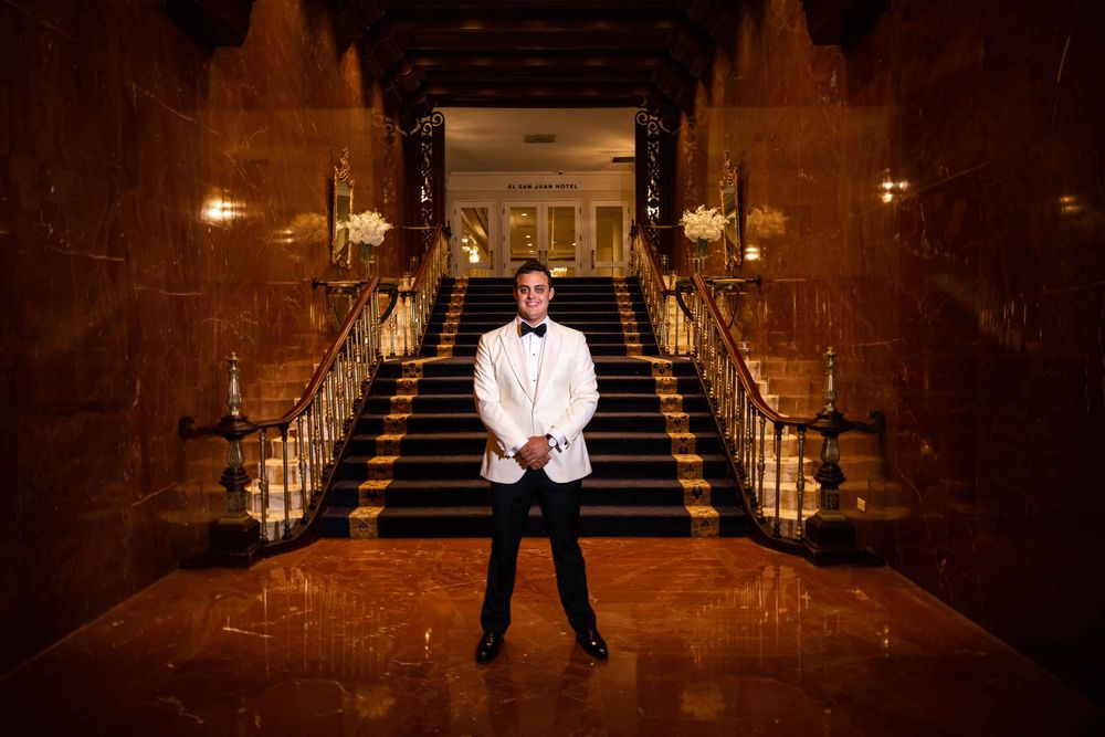 Someone in a white dinner jacket stands at the bottom of an elegant grand staircase with golden railings and warm lighting.