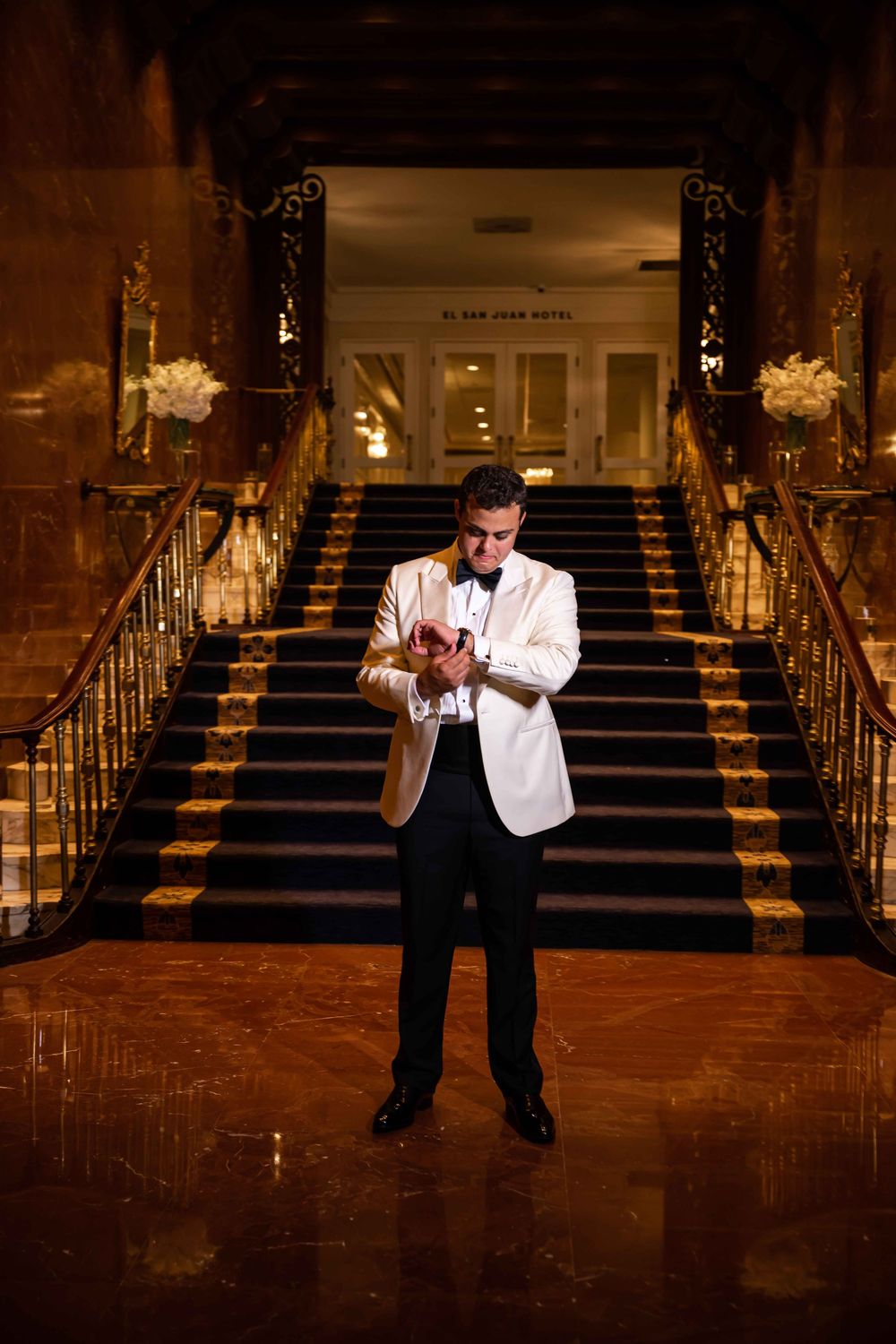 A person in a white dinner jacket checks their watch while standing on polished marble floors in front of an elegant staircase.
