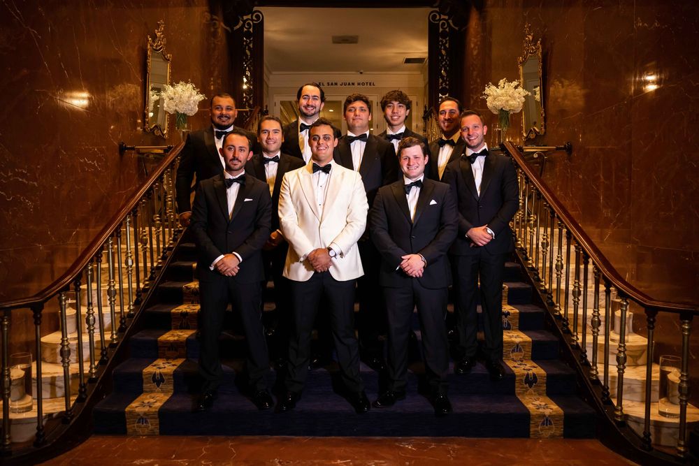 A group in formal black and white attire poses on an elegant staircase with ornate railings and warm lighting.