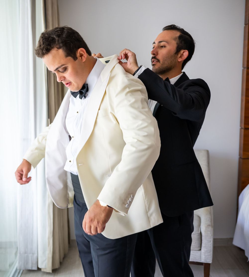 A groomsman helps adjust the white dinner jacket of another during wedding preparations beside a window.