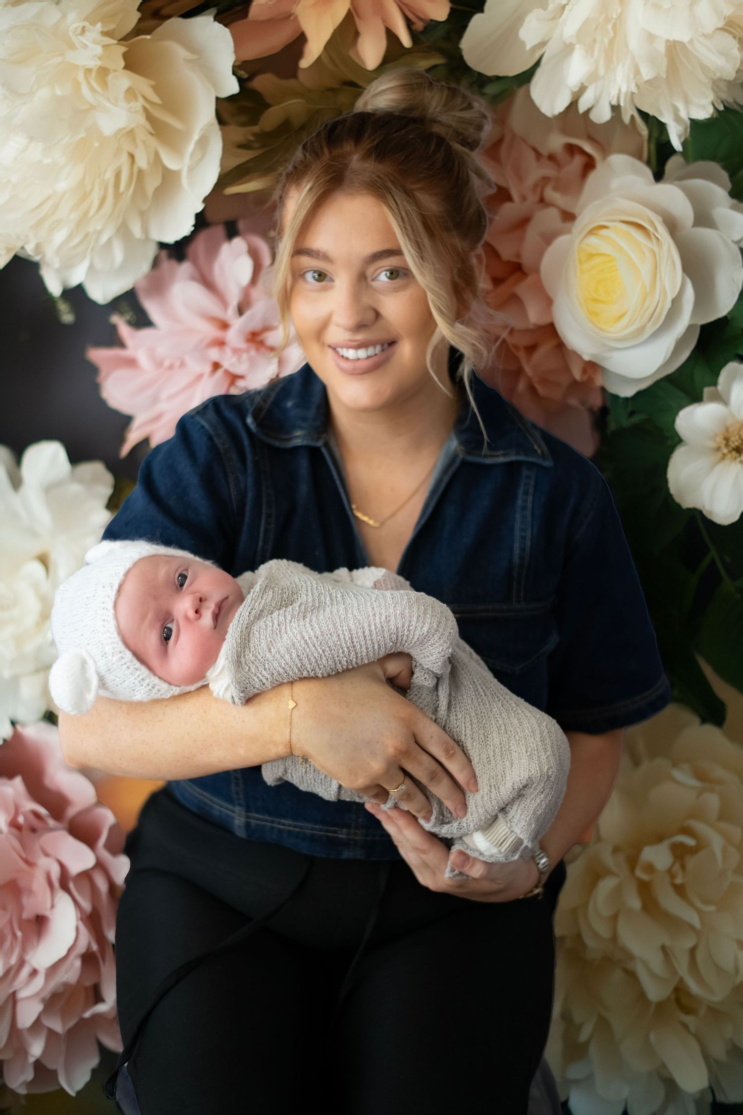 A nurse in dark scrubs holds a swaddled newborn baby against a romantic backdrop of pink and cream colored flowers.