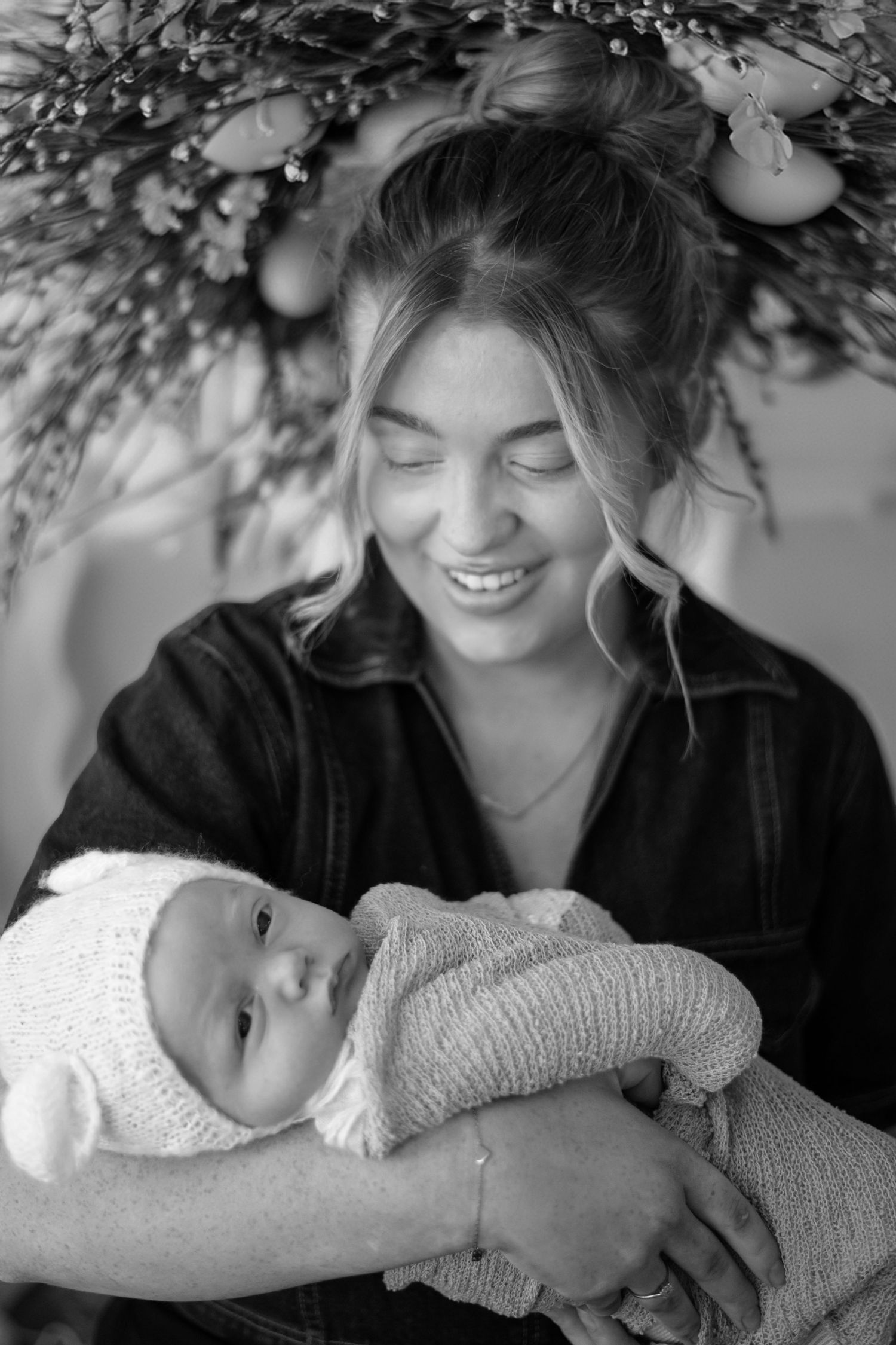 Black and white portrait of mother smiling lovingly while holding bundled newborn baby under Christmas decorations.