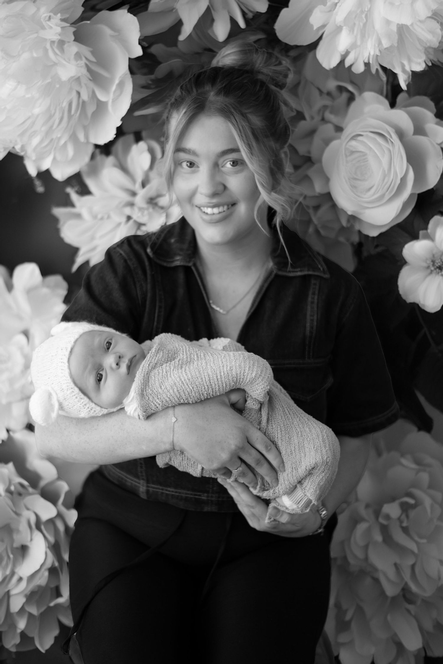 A black and white portrait shows someone in dark clothing holding a swaddled baby against a floral backdrop.