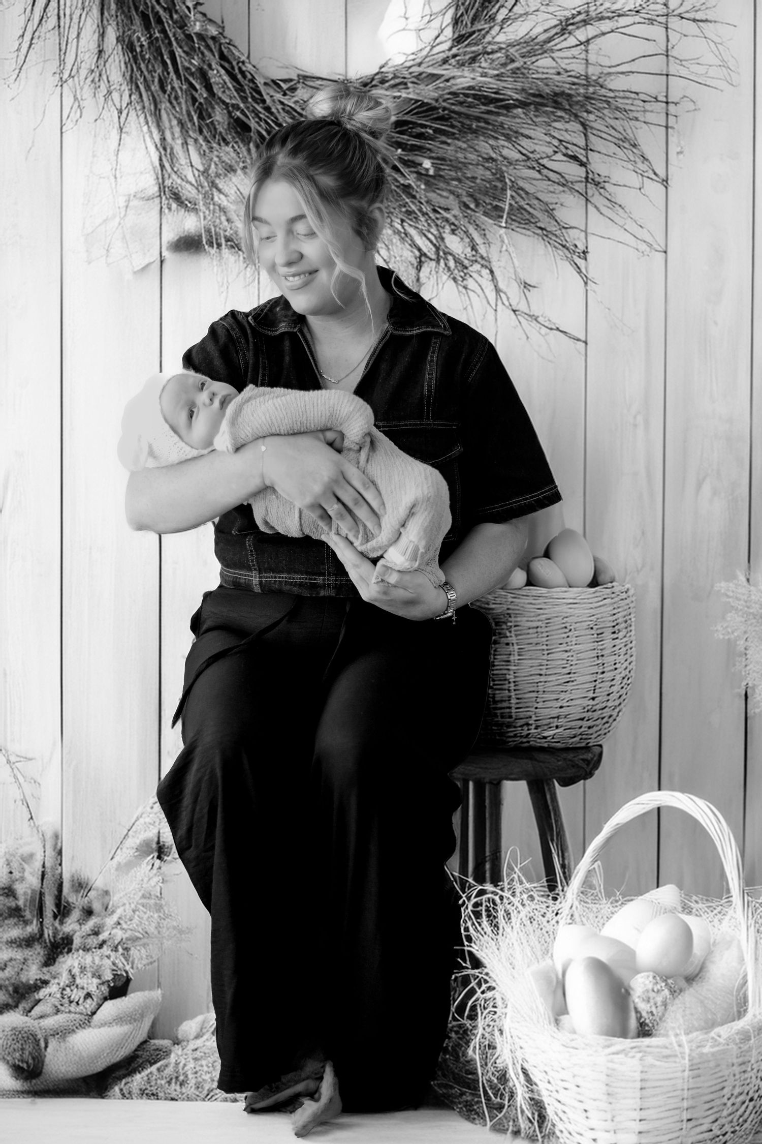 Black and white portrait of parent in dark clothing holding baby with decorative baskets and feathers in rustic setting.