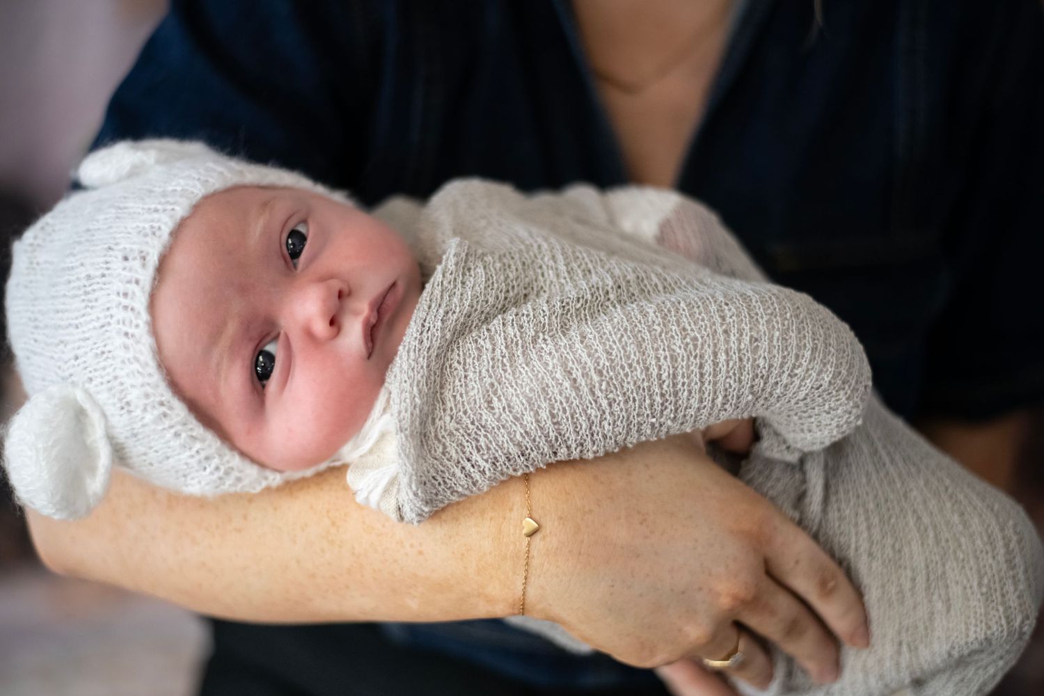 A newborn baby wearing a white knit hat and wrapped in a soft gray blanket rests in someone's arms.