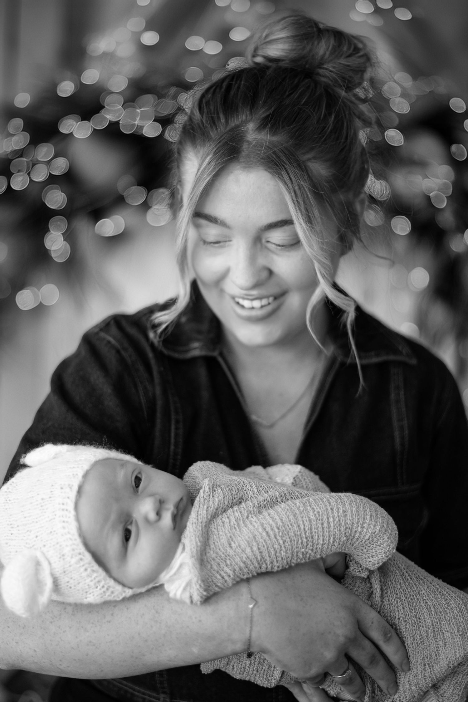 A heartwarming black and white portrait of a mother cradling her baby in a cozy sweater with twinkling lights in background.