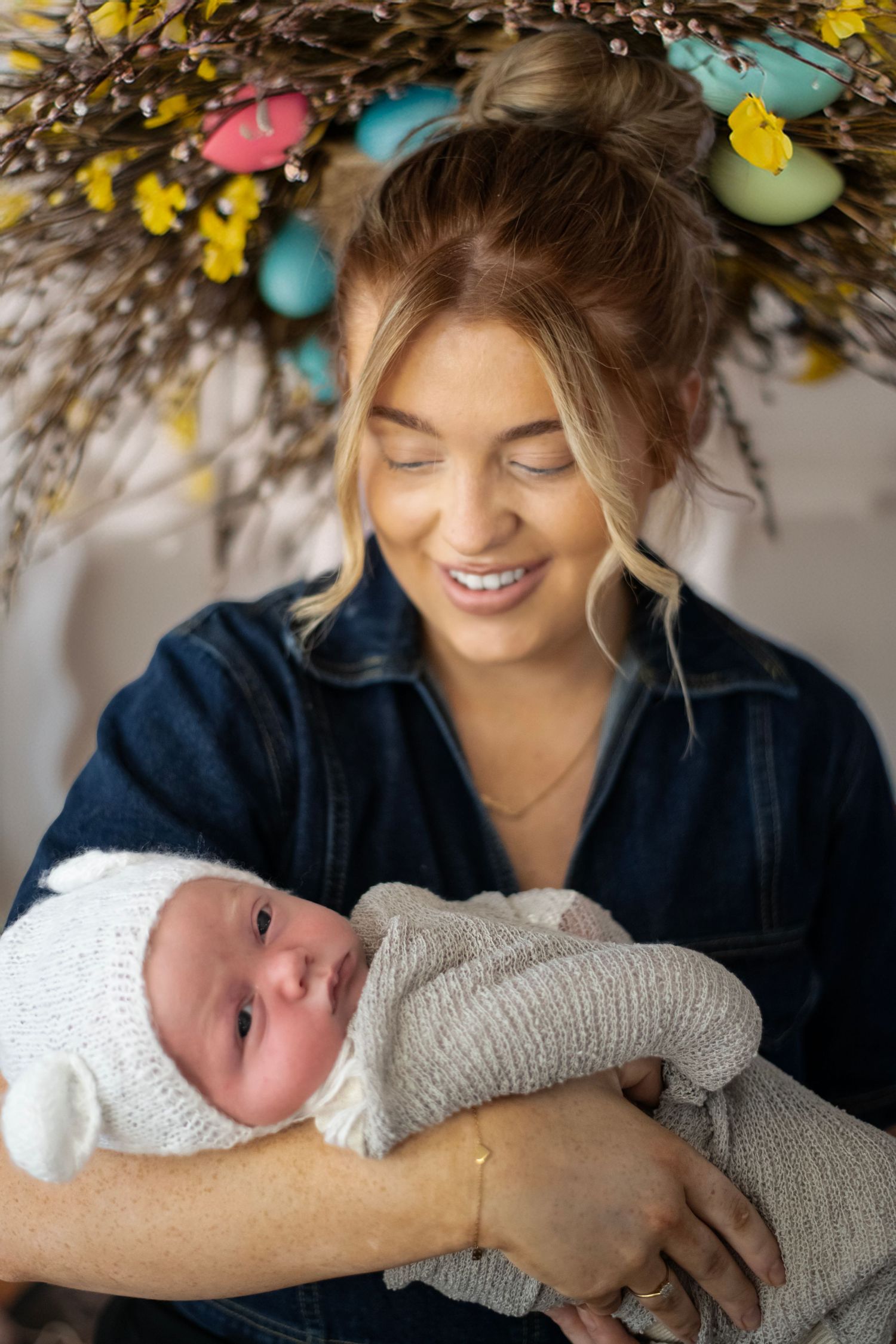 A mother holds her small newborn baby wrapped in a gray blanket near Easter decorations of colored eggs and spring branches.