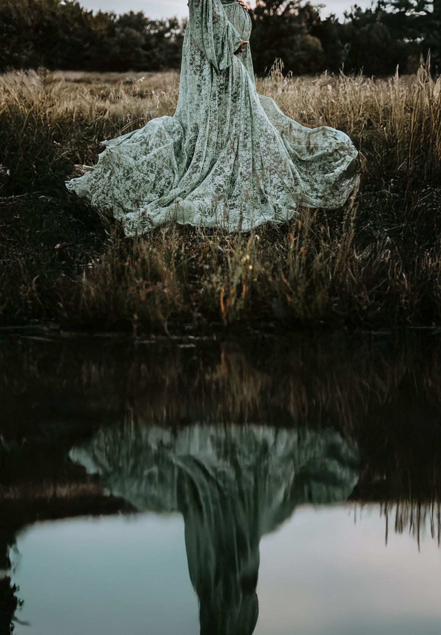 green lace maternity gown covers mother to be as she stands in a golden field at sunset next to the lake for her maternity photos