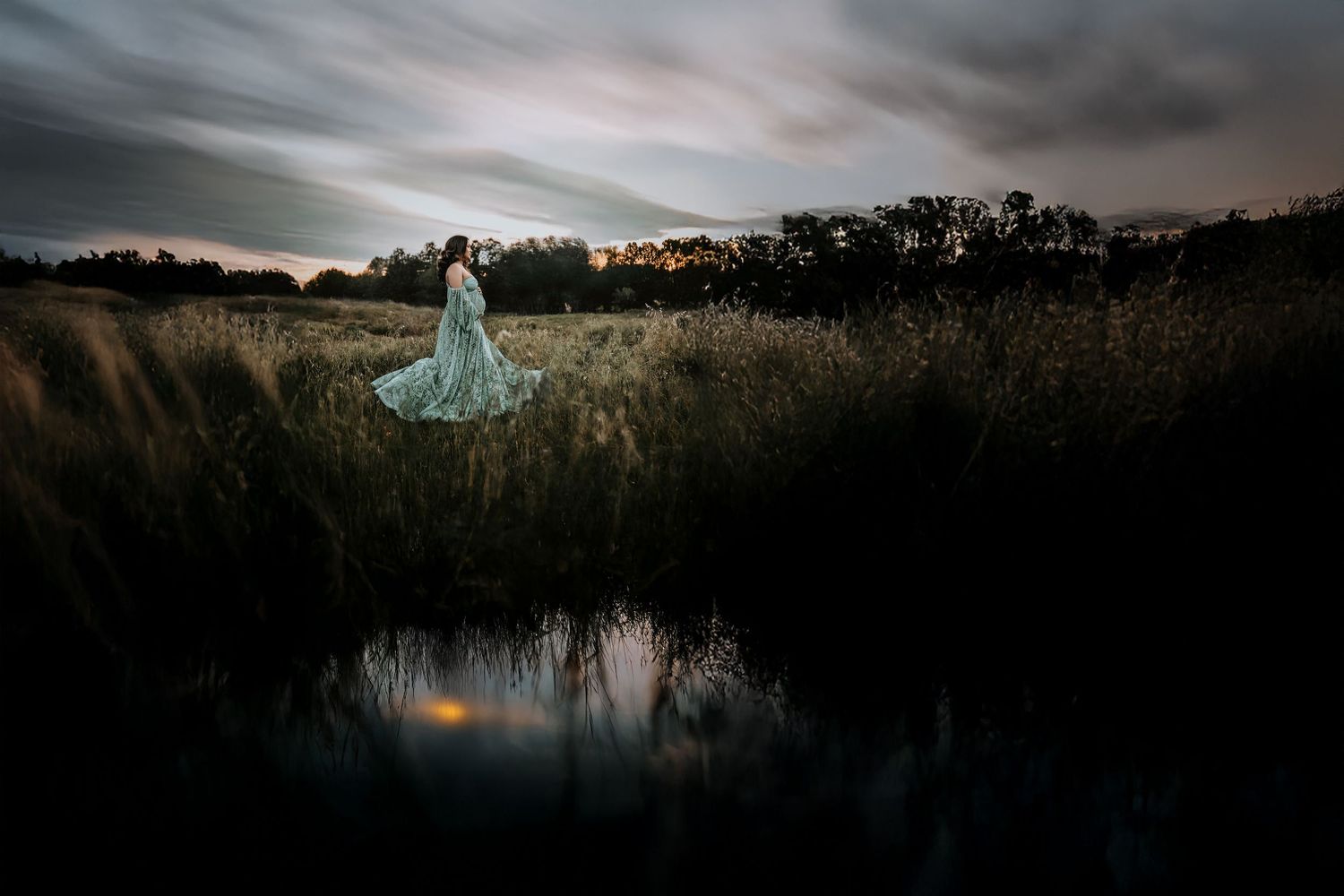 moody maternity photos of mother wearing lace mint green gown standing next to the lake with a dramatic sky