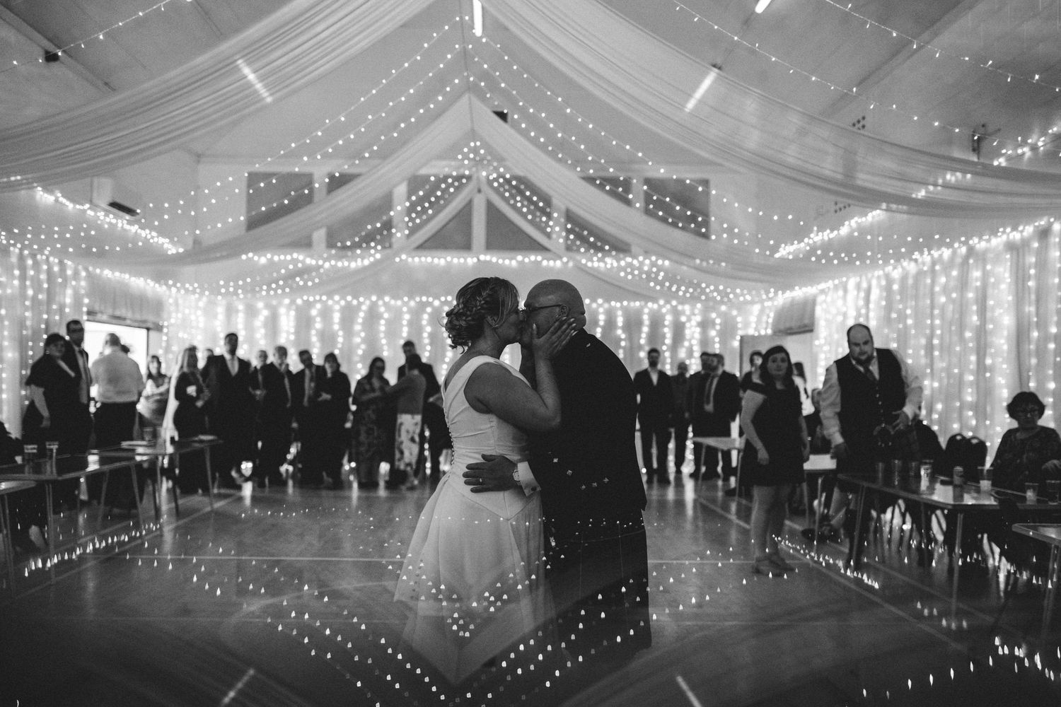 A couple shares an intimate dance moment in a beautifully lit reception venue with fairy lights and white draping.