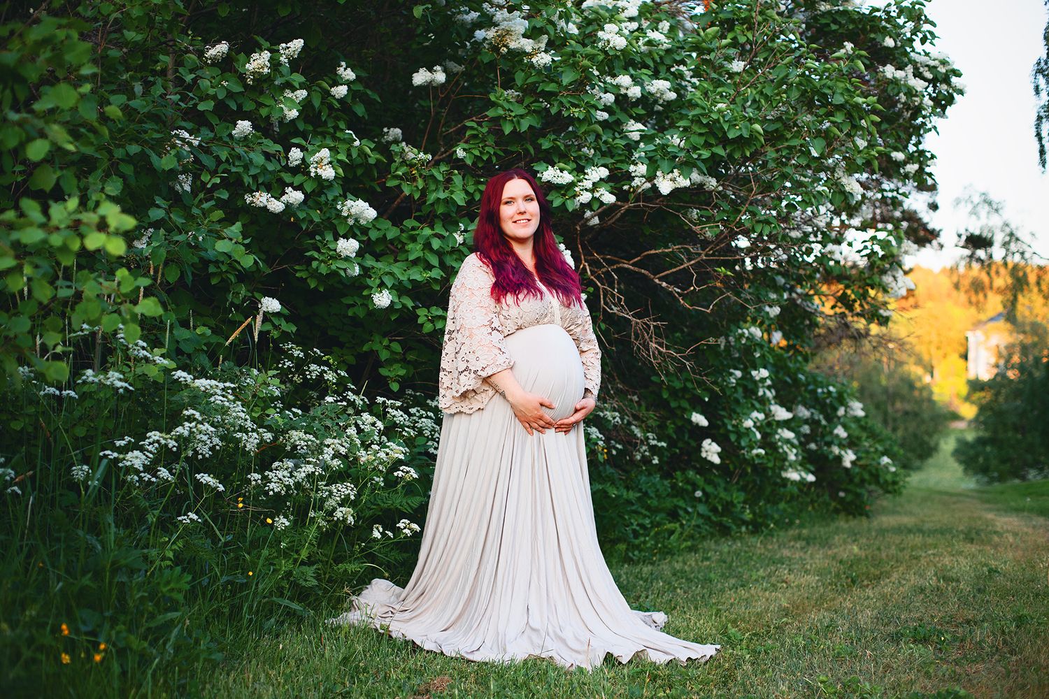 Pregnant woman in cream lace gown with red hair standing near blooming white shrubs.