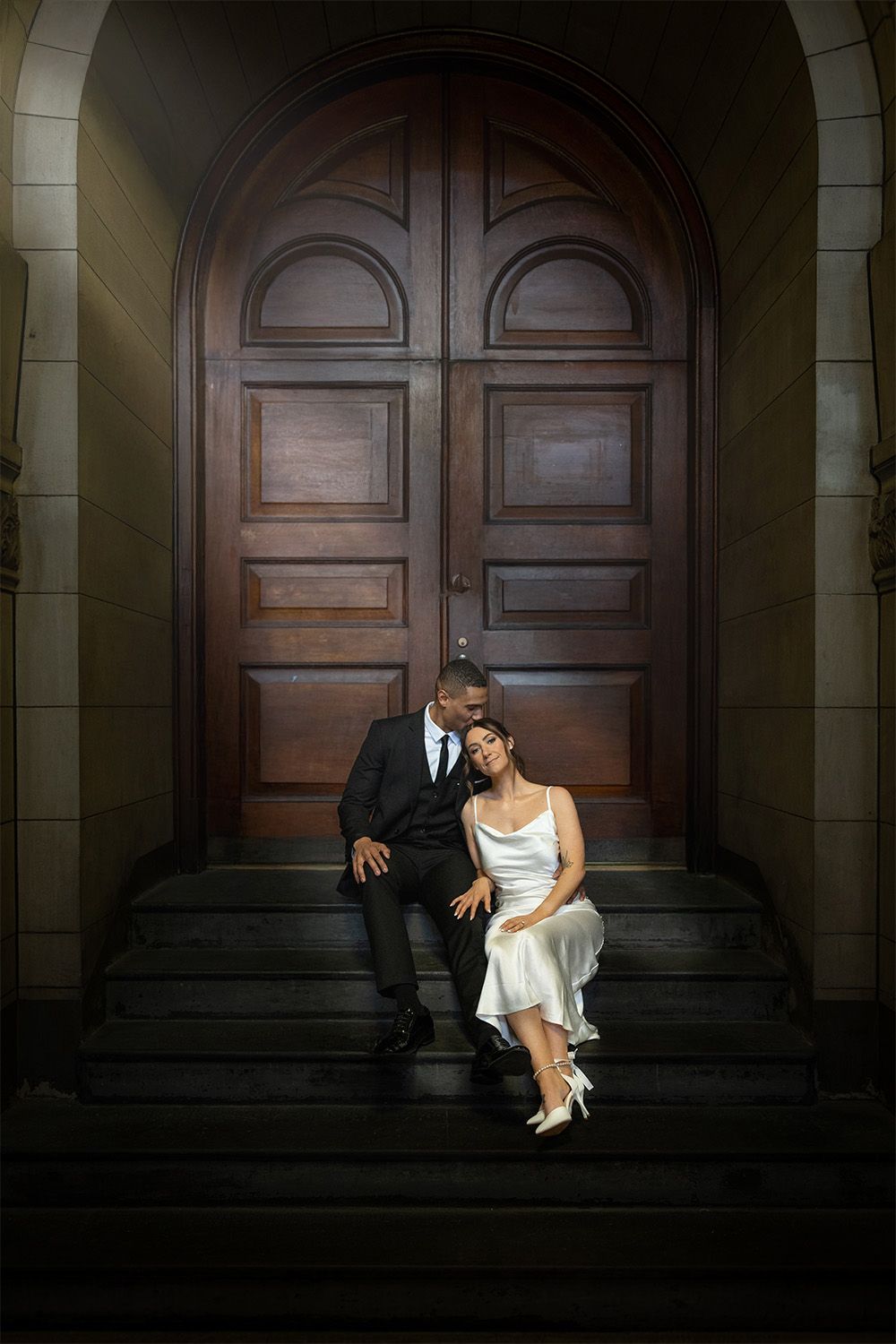 A couple in formal attire sits closely together on stone steps in front of large wooden double doors.