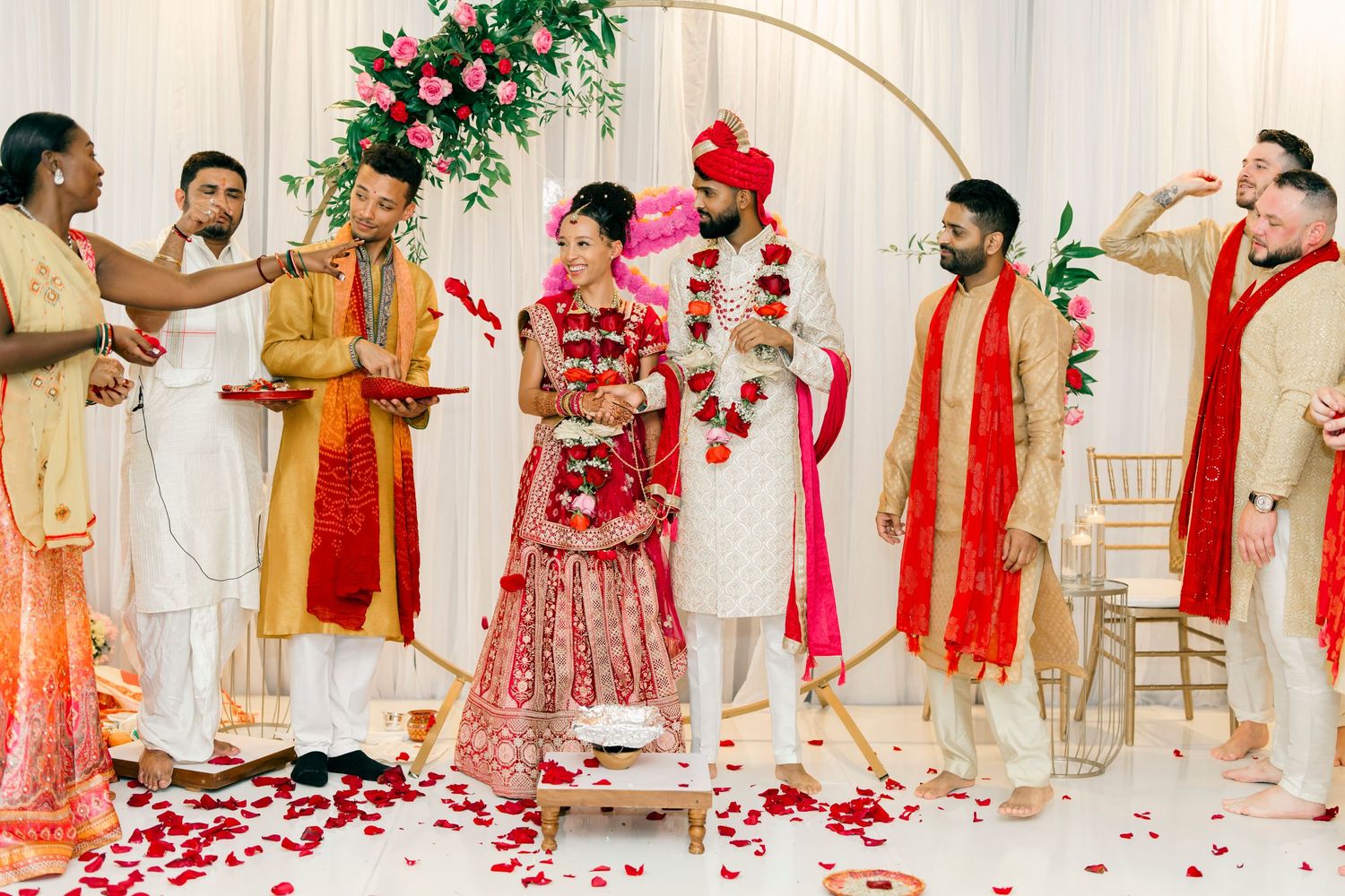 Traditional Indian wedding ceremony with people gathered around a floral arch decorated with white and red rose petals.
