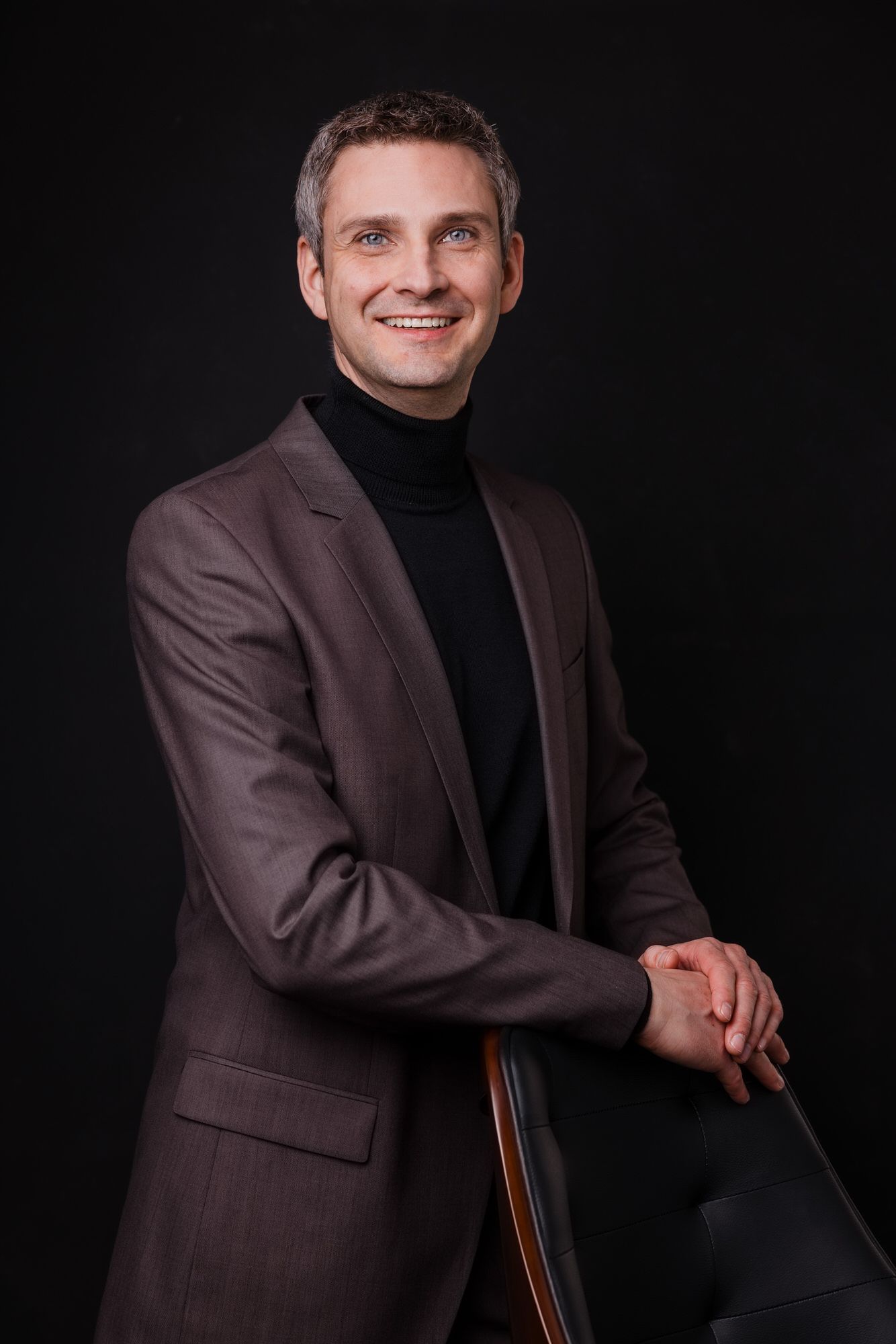 Professionelles Studioporträt einer Person mit braunem Blazer und schwarzem Rollkragenpullover vor dunklem Hintergrund.