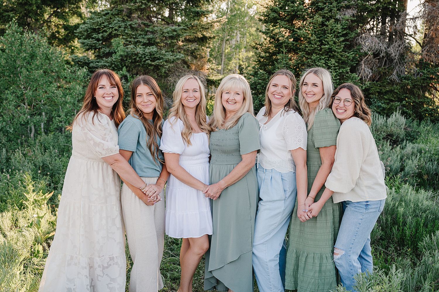 A group dressed in light pastel colors stands together outdoors in a natural setting with greenery and trees in background.