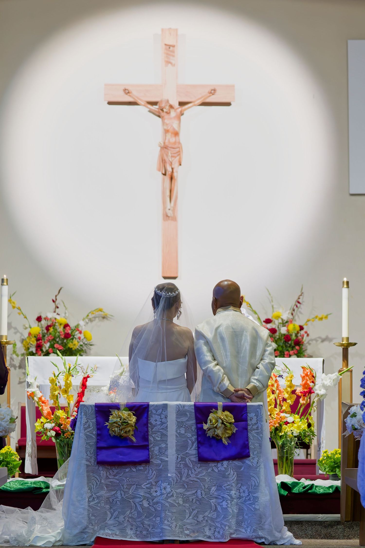 Couple kneels at church altar during wedding ceremony with crucifix and colorful floral arrangements in background.