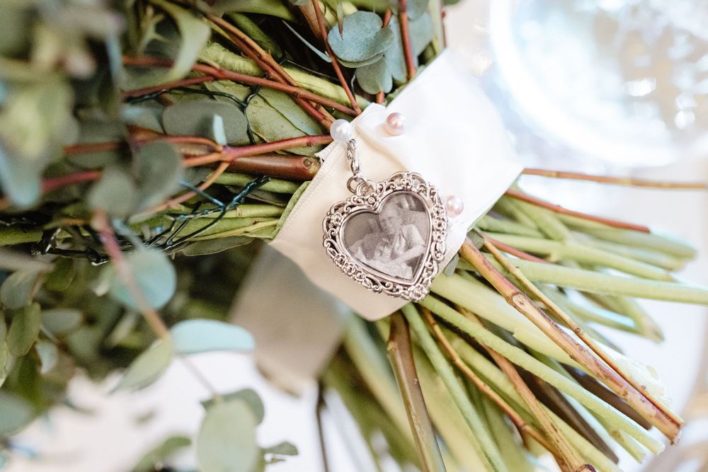 Wedding bouquet wrapped in white silk ribbon with an ornate silver crest brooch detail against a light background.