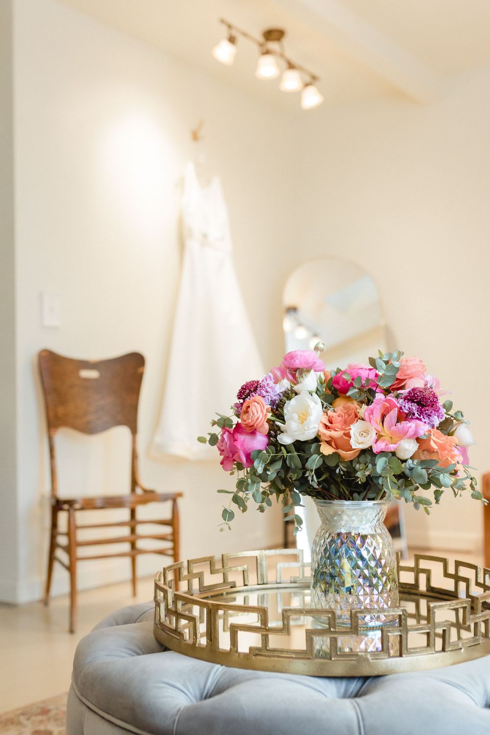 Vibrant pink and coral flower arrangement in crystal vase on gold geometric tray with wedding dress and chair in background.
