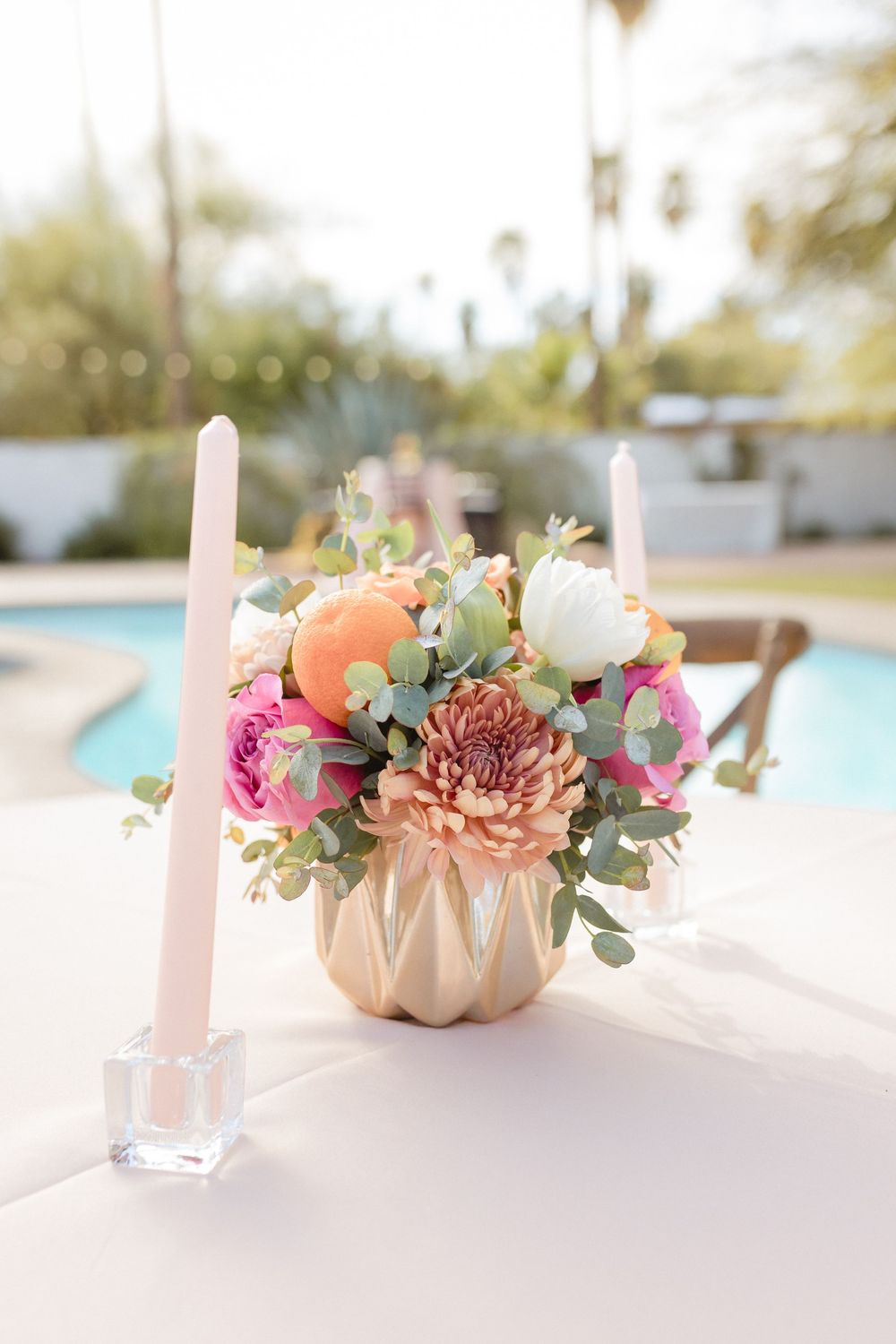 Romantic floral centerpiece with pink and peach blooms in a cream ceramic vase alongside white taper candles by a pool.
