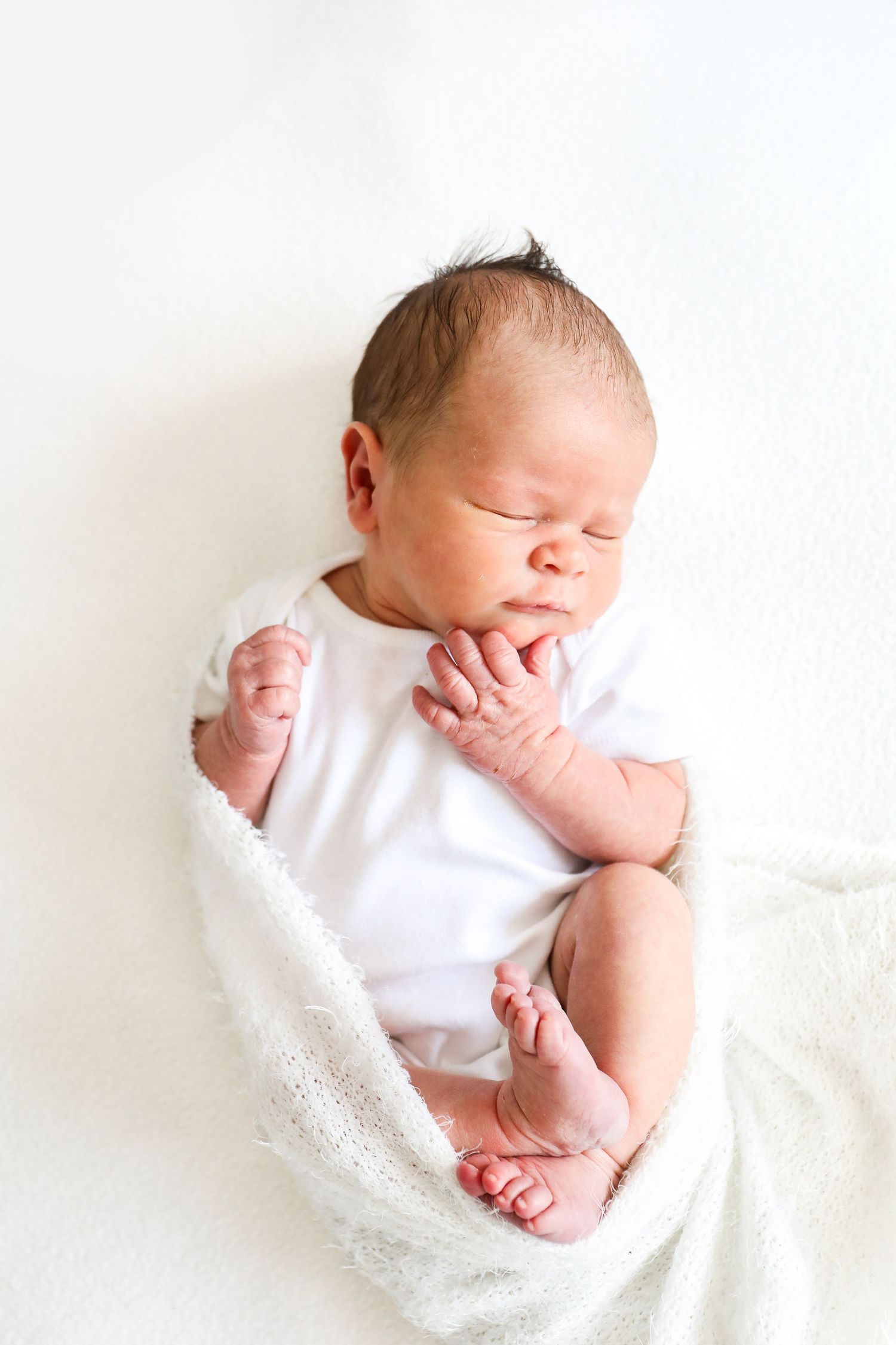 Sleeping newborn baby in white onesie lying on soft white blanket with hand near face, during newborn shoot in Nottingham