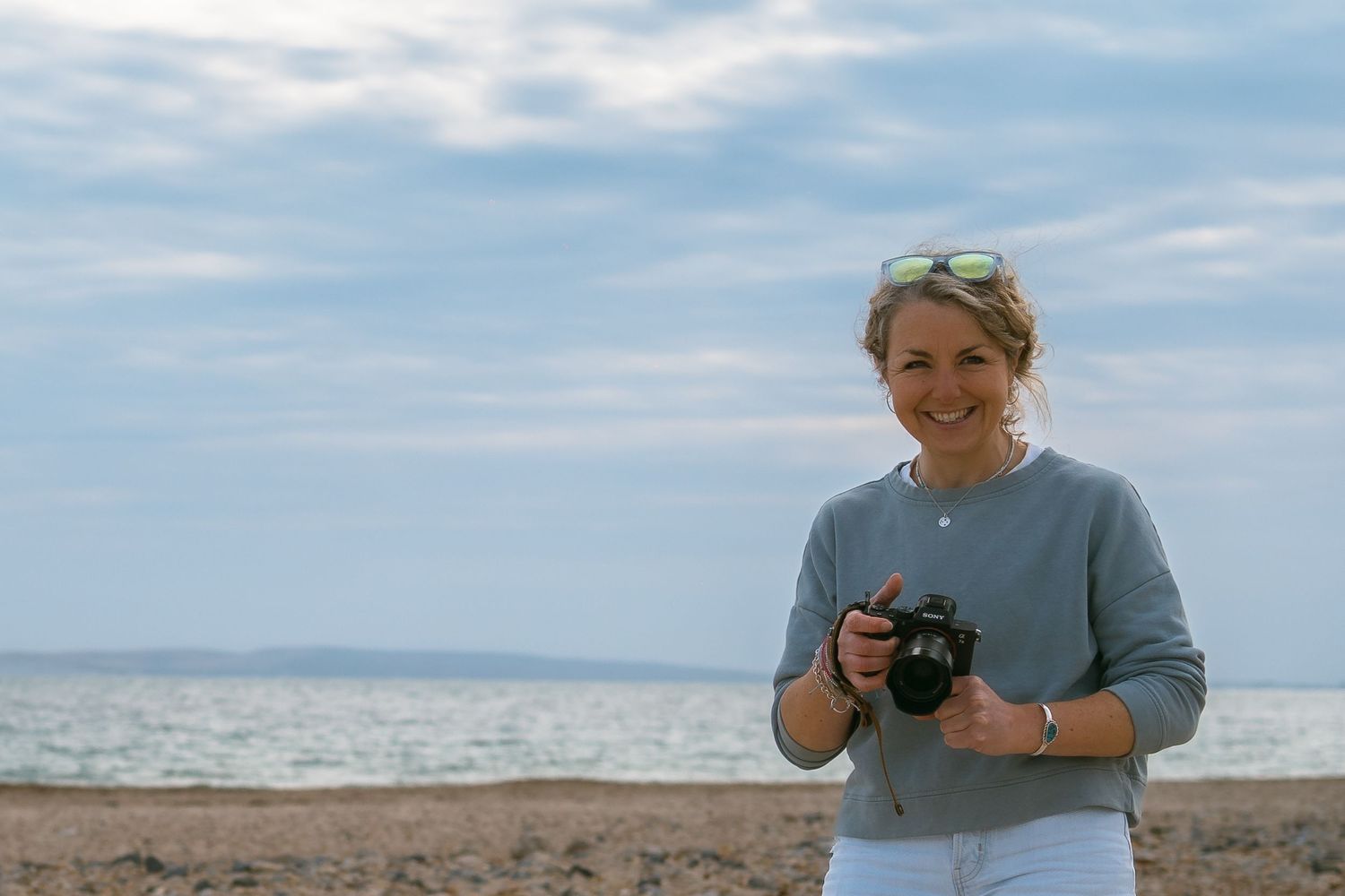 A selfie at a sandy beach with crashing waves and cloudy sky in the background, wearing a bright pink windbreaker.