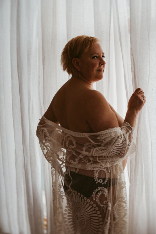 Woman in lace robe poses near white curtains in intimate boudoir portrait.