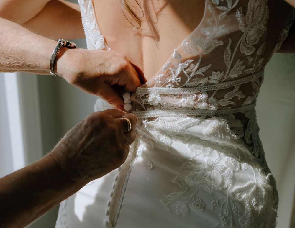 Close-up view of hands fastening buttons on the back of an ornate white lace wedding dress.
